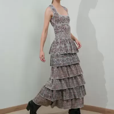 Mulher a usar vestido longo floral com camadas e botas pretas, em ambiente interior com parede cinza e chão de madeira.