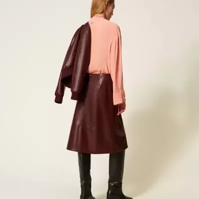 Mulher vista de trás com camisa rosa, saia de couro castanha, botas pretas e casaco castanho sobre o ombro