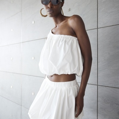 Mulher com roupa branca e acessórios dourados junto a parede cinza.