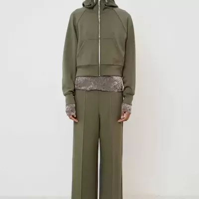 Conjunto verde-oliva com casaco de capuz e calças largas, top cinzento visível, e sapatos pretos de salto.