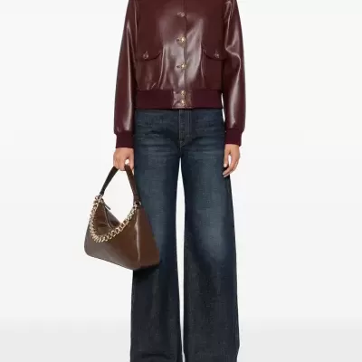 Mulher com casaco de couro castanho-avermelhado e calças de ganga largas, segurando mala castanha com corrente dourada