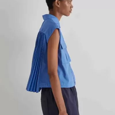 Blusa azul sem mangas com pregas atrás, combinada com calças azuis escuras em fundo neutro.