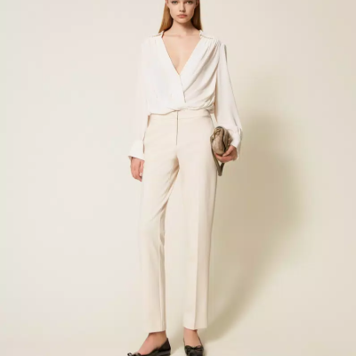 Mulher com blusa branca, calças claras e sapatos pretos num fundo creme