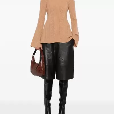 Mulher com camisola castanha, calções pretos de couro e botas altas pretas, segurando mala castanha com padrão cobra