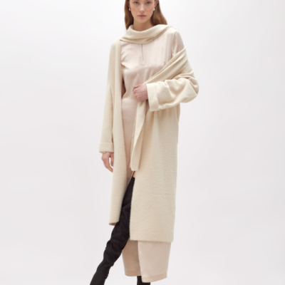 Mulher com casaco creme comprido, vestido cetim rosa claro e botas pretas em fundo branco