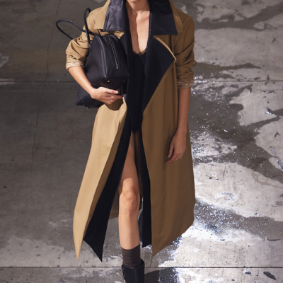 Mulher com casaco bege e forro preto, mochila preta, vestido preto, meias castanhas e botas pretas num chão industrial