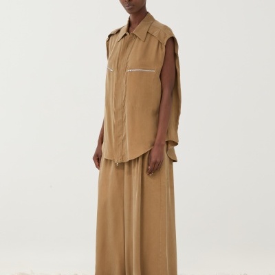 Modelo com conjunto castanho claro de camisa sem mangas com fechos e saia comprida, sobre chão de pedras brancas.