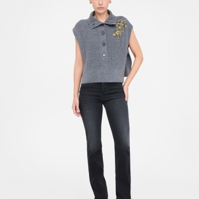 Mulher com camisola cinzenta de malha sem mangas com motivo floral amarelo no ombro, calças pretas e botas pretas