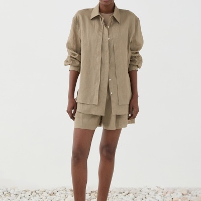 Mulher com camisa e calções bege claro, sapatos rasos castanhos, em chão com pedras brancas e fundo branco.