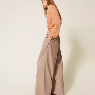 Mulher com calças largas bege e camisola de malha laranja posando em fundo claro