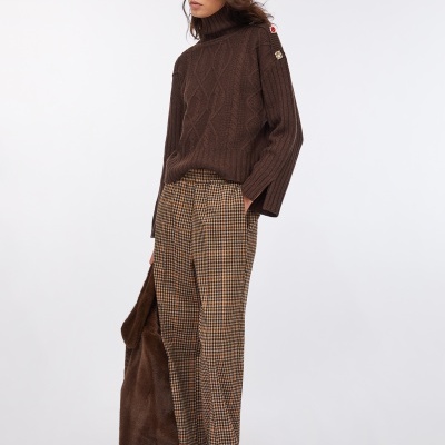 Mulher com camisola castanha de gola alta, calças xadrez castanho-bege, óculos escuros pretos e sapatos dourados, segurando casaco castanho de pelo