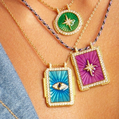Três colares com pendentes coloridos em azul, verde e violeta, adornados com pedras e detalhes em formato de estrela e olho.