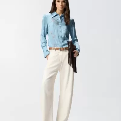 Modelo veste camisa azul acetinada com bordado floral, calças brancas largas, cinto bege, sandálias bege de salto e bolsa preta com franjas.