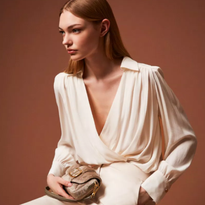 Mulher com blusa branca de tecido leve e calças claras segurando mini bolsa bege com fivela dourada