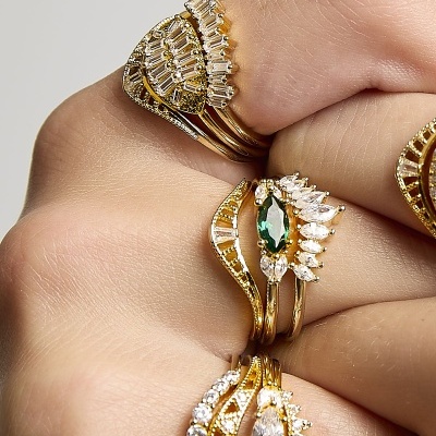 Mãos com vários anéis dourados com pedras brilhantes e detalhe verde.