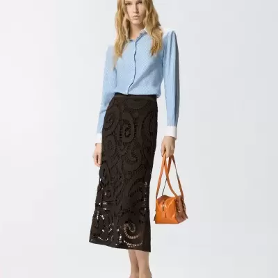 Mulher de camisa azul clara, saia midi preta em crochet, sapatos padrão pele de cobra e mala castanha em fundo branco.