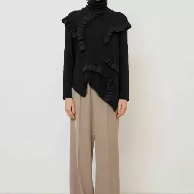 Mulher com camisola preta de gola alta com folhos e calças largas bege.