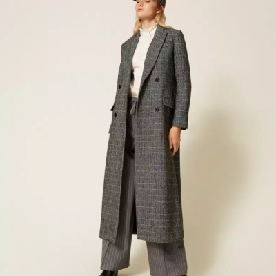 Mulher com casaco comprido cinza xadrez, camisa branca, calças riscas cinza, chapéu e sapatos pretos