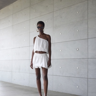 Mulher vestindo conjunto branco de top e saia curta com sandálias brancas e óculos de sol, em frente a uma parede cinza clara.