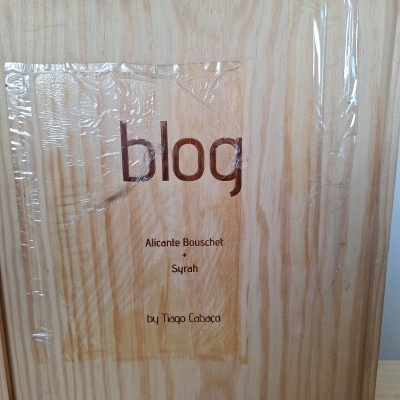 Caixa de madeira clara com texto 'blog Alicante Bouschet Syrah by Tiago Cabaço' em rótulo plástico