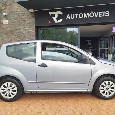 Carro prateado de três portas à frente de uma loja de automóveis com letreiro RC AUTOMÓVEIS e sinal OPEN