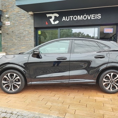 Carro preto ao lado de uma loja de automóveis com sinalização 'RC AUTOMÓVEIS' e placa OPEN neon