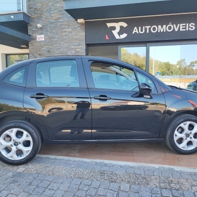 Carro compacto preto de lado junto a um edifício com placa RC Automóveis.