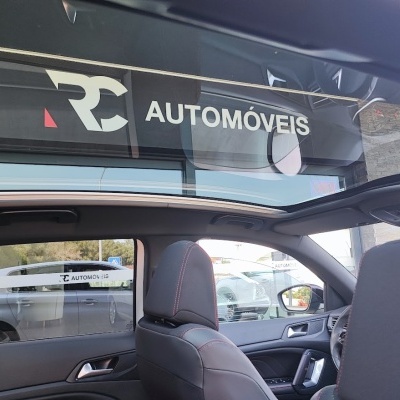 Interior de carro com bancos em couro preto e teto panorâmico aberto num stand de automóveis com letreiro 'RC AUTOMÓVEIS'