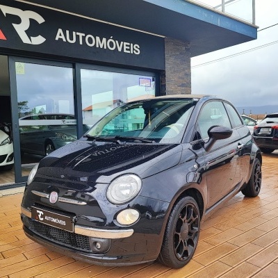 Carro Fiat 500 preto com logotipo RC Automóveis à frente de loja com outras viaturas estacionadas.