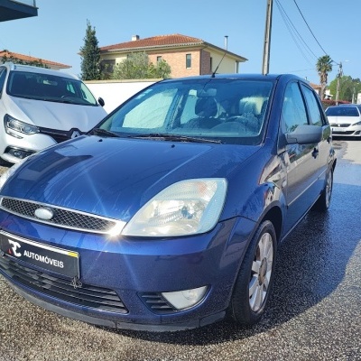 Carro Ford azul molhado com placa AUTOMÓVEIS estacionado numa estrada com outro carro branco atrás e casas ao fundo