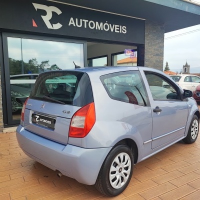 Carro Citroën C2 cinza prateado junto a loja RC AUTOMÓVEIS em pavimento de madeira