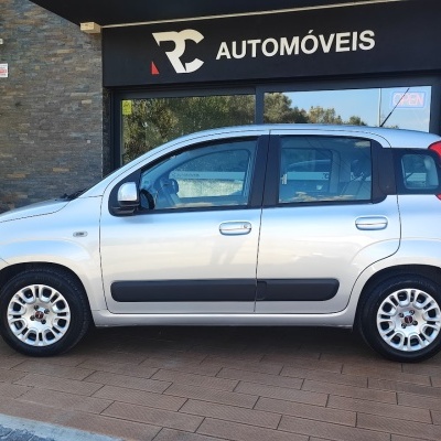Carro prata estacionado junto a loja RC Automóveis com letreiro e sinal de neon aberto