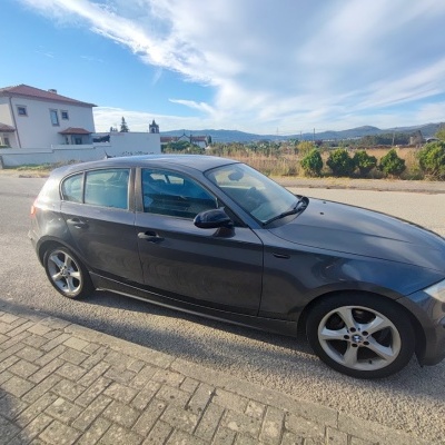 Carro BMW cinzento estacionado numa estrada com casas ao fundo.