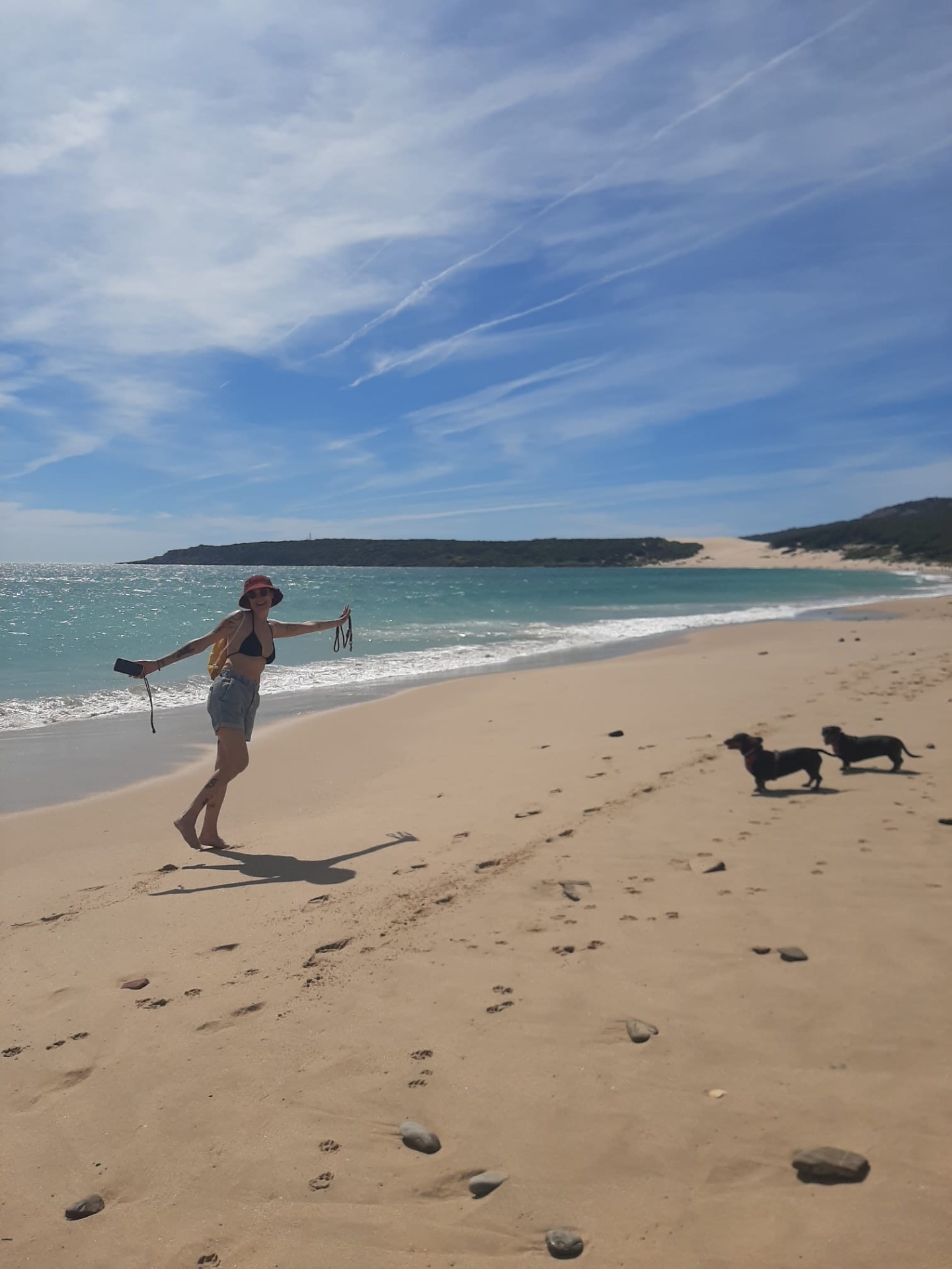 🐾 As Aventuras da Piaf & Farofa - Playa de Bolonia - Cadiz/ES - Por Tia Carla | Real Pet Grooming Praia com mulher e dois cães na areia junto ao oceano sob céu azul com nuvens.