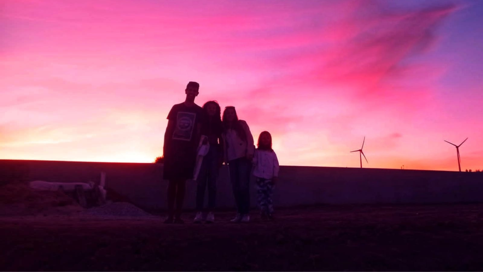 Silhuetas de quatro pessoas ao pôr do sol com turbinas eólicas ao fundo