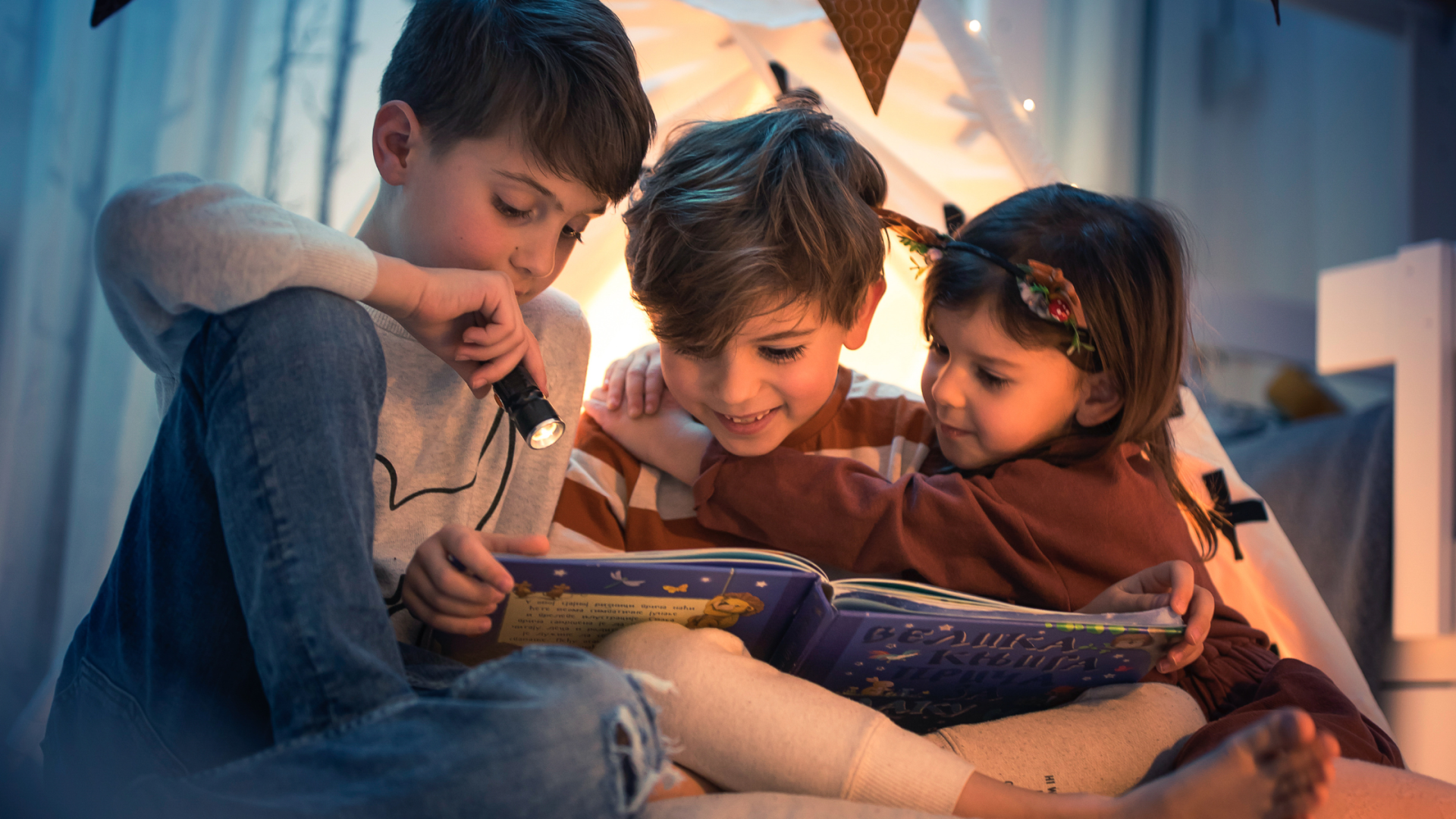 Três crianças a ler um livro infantil com uma lanterna dentro de uma tenda decorada com luzes
