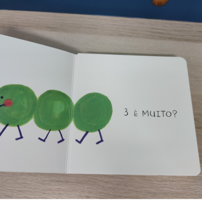 Livro infantil aberto com três círculos verdes e texto '3 É MUITO?'