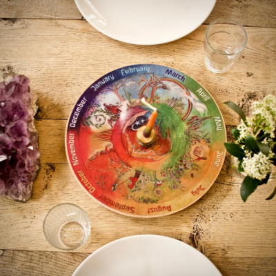 Relógio colorido com meses do ano sobre mesa de madeira com pratos, copos, cristal e planta