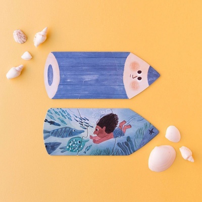 Dois marcadores de livros de madeira em formato de lápis com ilustrações, sobre fundo laranja, rodeados por conchas brancas.