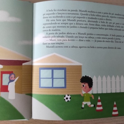 Página de livro infantil com ilustração de criança a jogar futebol na relva entre casas e cones.