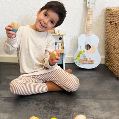 Menino sentado no chão com pijama às riscas segurando ovos de madeira, guitarra branca infantil e cesta de vime ao fundo