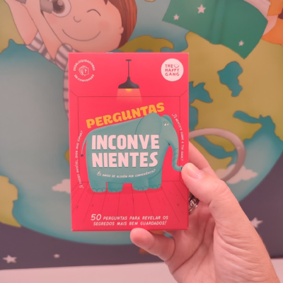 Caixa vermelha de cartas 'Perguntas Inconvenientes' com elefante azul e texto, mão humana segurando, fundo colorido desfocado.