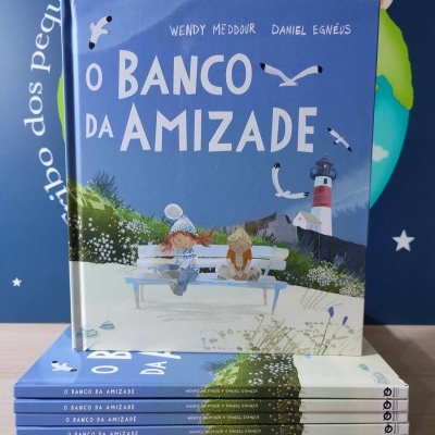 Capa do livro 'O BANCO DA AMIZADE' com crianças num banco, fundo azul com farol e gaivotas, seis livros empilhados sobre mesa