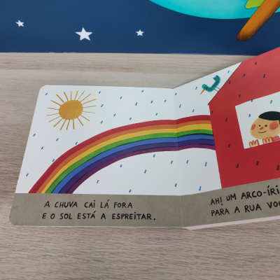 Livro infantil aberto com arco-íris, sol, chuva e casa vermelha