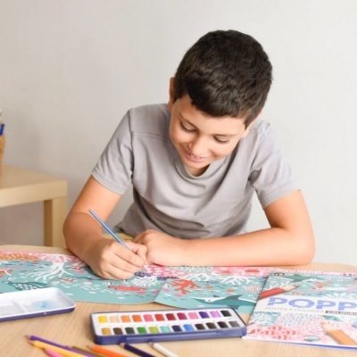 Menino pintando desenho floral com aguarelas numa mesa com materiais de pintura