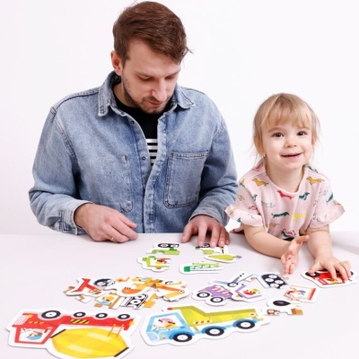 Adulto e criança a montar puzzle de madeira de veículos coloridos numa mesa branca