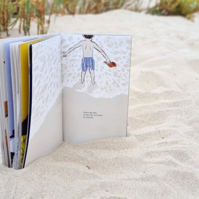 Livro aberto na areia da praia com texto em inglês e ilustração de pessoa a segurar balde vermelho