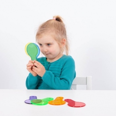 Criança segurando espelhos coloridos sentada à mesa branca