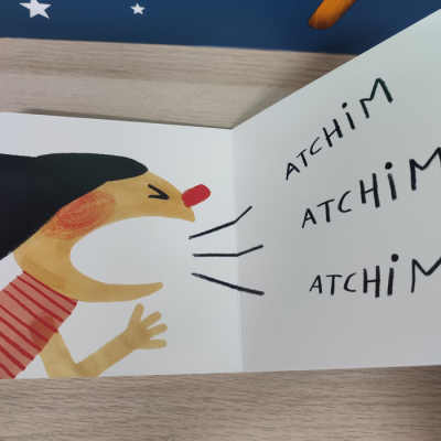 Ilustração de personagem a espirrar num livro infantil aberto com texto 'ATCHIM' repetido.