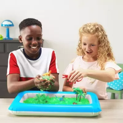 Crianças brincando com brinquedo de dinossauros na areia verde dentro de piscina insuflável azul e branca